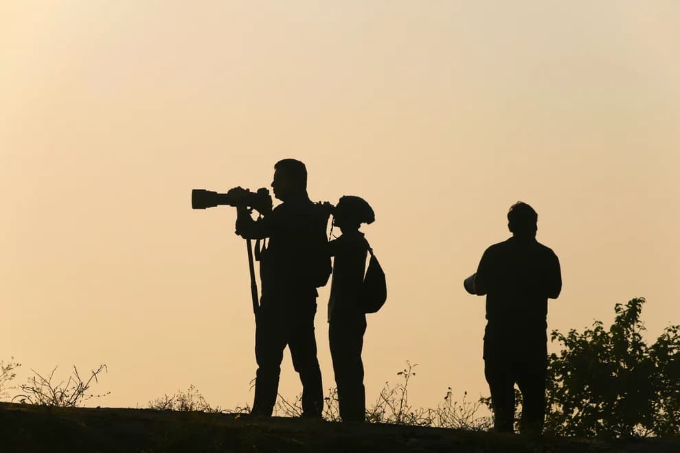 Three silhouetted birders with binoculars and a camera at golden hour